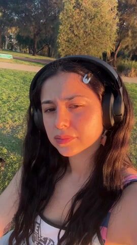 A woman in a park is listening to something through headphones. She looks concerned and is speaking. The woman has long dark hair and is wearing a white top with a design on it. She also has earrings and a hairpin. The park has green grass and some trees. The sun is shining on the woman's face.