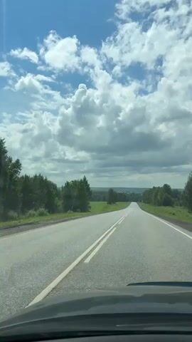 The media file shows a POV shot from inside a vehicle driving on a paved road. The road is asphalt and has white dividing lines. The surrounding environment is rural, with green fields and trees on either side of the road. In the distance, the road appears to curve and disappear over a gentle hill. The sky is a bright blue with scattered white, fluffy clouds, suggesting a sunny day with some cloud cover. A bright sun is visible, casting lens flares in the upper left portion of the frame. The time of day appears to be daytime, likely late morning or afternoon, given the brightness of the sun. The location is identified as Shlyapniki, Russia. There is no visible text within the media file. The overall mood is one of travel and openness, typical of a scenic drive through the countryside. The scene is devoid of people or other vehicles, emphasizing the feeling of a solitary journey.