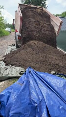 A red dump truck is captured in an outdoor setting, actively unloading a large quantity of dark brown material, likely soil or compost. The truck's bed is fully tilted, and the material is cascading onto two large tarps spread on the ground: a green one directly beneath the pile and a blue one folded in the foreground. In the background, a dirt road or path is visible, with a white car parked further down. To the left, there are trees and foliage, and to the right, part of a building with a green fence can be seen. The sky is bright with some clouds, indicating daytime. The truck's rear license plate displays "980 6754", and a circular "80" sticker is also visible, likely indicating a speed limit for the vehicle. The scene appears to be taking place in Nový Jáchymov, Czechia, as suggested by the license plate.