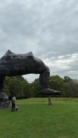 A man and a child walk across a grassy field towards a large, dark-colored sculpture resembling an inverted human figure with legs bent, supporting the torso. The sculpture is set against a backdrop of green trees and a cloudy sky. The figure's head is positioned upside down on the ground. The man is wearing a backpack, and the child is a toddler in a pink outfit. The sculpture appears to be made of metal and has a textured surface. The scene is outdoors, possibly in a park or sculpture garden.