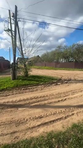 The camera pans slowly across a dirt road in a rural or suburban area on a partly cloudy day. The road is sandy and rutted, indicating vehicle traffic. Power lines run overhead, supported by wooden poles. On either side of the road is grass and vegetation, bordering residential properties with fences. Houses are visible behind the fences, typical of a countryside or suburban setting. The lighting is bright, suggesting a sunny day despite the cloud cover. No people or animals are visible in the frame.