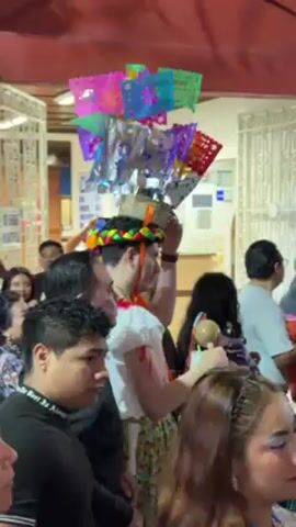 A person dressed in traditional Mexican clothing, including a floral head wreath, a white embroidered blouse, and a colorful skirt, stands in a crowd of people under a covered walkway. The person also has a tall decorative structure on their head with colorful paper cutouts. Most of the people around them are looking at them and some are filming with their phones. A few people in the crowd are also wearing similar floral wreaths. The walkway has a red roof and the crowd is gathering outside a building with a sign that reads "CIRO." The music is lively and upbeat, suggesting a festive atmosphere.