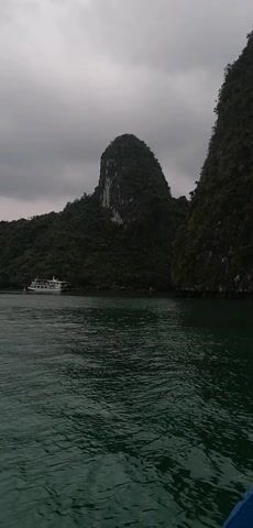 The video presents a scenic view of Ha Long Bay in Vietnam on an overcast day. The camera captures a wide shot of the bay featuring striking karst formations covered in dense green vegetation rising from the dark green water. A white cruise boat sails across the water. The sky is filled with gray clouds, casting a soft, diffused light on the landscape.  The water is calm with gentle ripples. The focus is on the natural beauty of the bay.