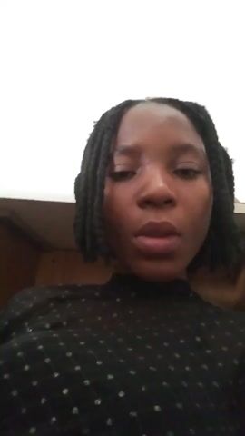 A young woman with dark braided hair is speaking to the camera. She is wearing a black top with a subtle pattern. The background is blurred, but appears to be indoors. Her right hand is raised to her face, touching her chin at one point.