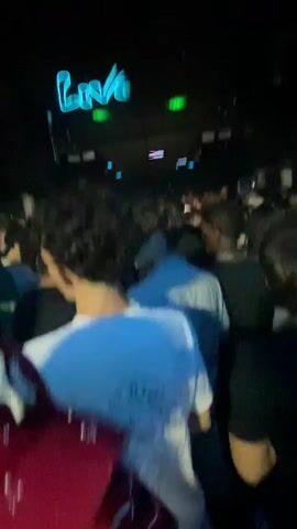A video taken from within a crowded, dimly lit nightclub shows a view of the dance floor and the DJ booth area. Neon lights illuminate the space, with a prominent neon sign reading "Liva" visible above the DJ booth. People are packed together, facing the DJ booth, with some raising their hands. The lighting shifts between blue and green hues, and spotlights are directed at the crowd. The perspective is from someone standing in the crowd, looking towards the DJ.