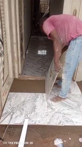 The video shows a person in a pink t-shirt and blue jeans installing white and gray marble tiles at the entrance of a building. The person is working barefoot, using a trowel to spread mortar on the ground before placing the tile. Some tiles have already been laid down. A level is on the ground, near the newly laid tiles. The doorway to the building is visible behind the worker, and it has marble tile already installed. The video is taken from a low angle, showing the person's feet and the ground. A "TECNO SPARK 20" watermark is visible at the bottom left corner of the frame.