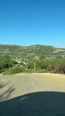 The video shows a landscape with rolling hills covered in green vegetation, likely farms or orchards, under a clear blue sky. The perspective appears to be from a vehicle moving along a dirt road or path, with a portion of the vehicle's interior visible in the foreground. A guardrail or barrier is also present in the lower part of the frame. The camera is panning or moving, offering a view of the expansive agricultural scenery.