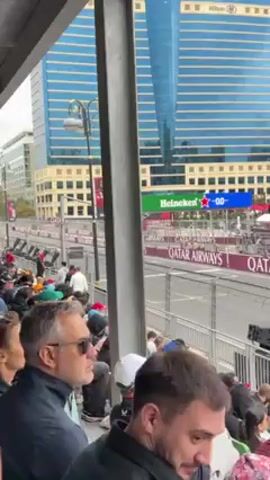 From a seated position in a spectator stand, the video captures a Formula One race in an urban setting. Two race cars, one white and red and the other orange and blue, speed along a straightaway track, passing in front of a grandstand filled with spectators. Large Qatar Airways signage lines the track. In the background, a tall blue glass skyscraper is visible, identified by a small "Hilton" logo near the top. Other buildings dot the cityscape. The sky is overcast. As the cars pass, the camera pans slightly to the left, following their movement and revealing more of the city's buildings. The video captures the noise of the race.