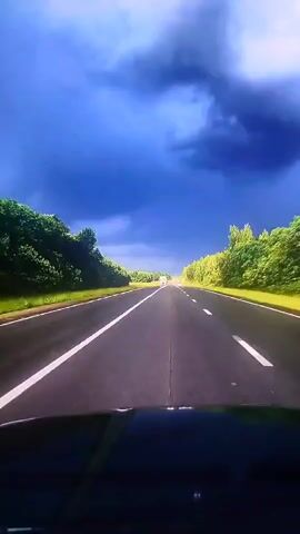 A first-person perspective shot from inside a vehicle shows a dark, stormy sky with heavy, dramatic clouds overhead. The vehicle is traveling on an empty, two-lane highway that curves slightly to the right in the distance. Lush green trees line both sides of the road, and the sun is visible in the far distance, creating a bright spot. A white semi-trailer truck is seen in the middle distance, and a few other vehicles are also present on the highway. The camera is focused on the road ahead, with the dashboard of the vehicle visible in the foreground.