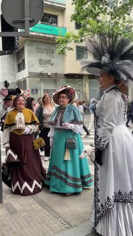 The video shows three women standing on a city street dressed in what appears to be Victorian-era costumes. The woman on the left is wearing a brown dress with a tan capelet. The woman in the middle is wearing a turquoise dress with a matching capelet. Both are wearing large brimmed hats adorned with flowers, and they're carrying purses. The woman on the right is wearing a long white coat-dress with black accents and a large hat with black feathers. A white dog is partially visible behind the woman in the middle. There are other people walking in the background, and a photographer is taking pictures of the women. A street sign and the façade of a building, labeled "dorsia Eva Cliniques" are visible in the background. The weather appears to be pleasant and sunny.