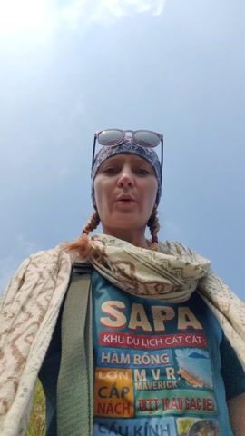 A woman with reddish-blonde hair in braids is recording a selfie video. She is wearing sunglasses on her head, a patterned bandana, a patterned scarf, and a teal t-shirt with images and Vietnamese writing on it. She also wears a patterned overshirt and a green crossbody bag, and has a plaid garment tied around her waist. She is standing on a cliff or mountainside, with a view of a lake, mountains, and mist in the background. She turns to show the view and smiles.