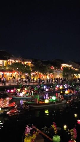 The video shows a nighttime view of a waterfront area in a city. Several boats are floating in a dark river, each lit with colorful glowing orbs. People are seated in the boats, and many of them are visible on the bank as well. Buildings along the bank are illuminated with traditional red lanterns and other lighting, creating a warm and festive atmosphere. The sky above is dark and clear. The water reflects the lights from the boats and buildings.