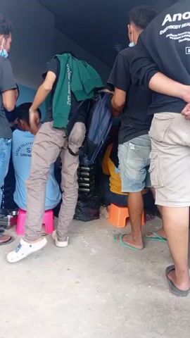 A group of young men and women are gathered in an outdoor area. One man on the left, wearing a black t-shirt, blue jeans, and a face mask, looks towards the center. To his right, a woman in a blue headscarf and blue shirt sits on a pink stool. Next to her, a man in a black t-shirt and grey pants stands with a green hoodie draped over his shoulders and a blue backpack slung over his arm. In the center, a woman with a black hijab and a yellow shirt sits on an orange stool, looking down. To her right, a man in a black hoodie and khaki shorts stands with his back to the camera, holding a cigarette. Another person is partially visible on the far right, wearing a black hoodie and khaki shorts. The ground is concrete, and a blue wall is visible in the background. Objects include several small plastic stools, bags, and what appear to be stacked metal items. The overall impression is a casual gathering, possibly waiting for something or someone.