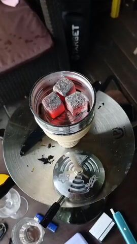 The media file shows a close-up, top-down view of a hookah bowl filled with glowing red charcoal cubes. The hookah is placed on a metallic tray with a heat management device, also metallic and engraved with a logo that appears to read "RAZZOR". The charcoal is actively burning, emitting a faint glow and some ash.

The setting appears to be outdoors, possibly a patio or deck, with wooden planks visible in the background. There are some blurry elements suggesting furniture, possibly a wicker chair with a pink cushion, and a black bag with the word "TARGET" printed on it. In the foreground, on the table, there are a few glasses, a lighter, and a small blue electronic device, perhaps a vape. The lighting suggests it might be daytime, though the exact time is unclear. The overall impression is a relaxed, casual atmosphere, likely a moment of preparing or enjoying a hookah session. No people are visible in the frame.