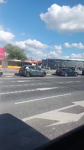The media file depicts a street scene in Mysiadło, Poland, on a sunny day with scattered clouds. The camera is positioned inside a vehicle, likely a bus or van, looking out at the road.

The primary subjects are vehicles on a multi-lane road. In the foreground, the edge of a red car is visible. Further down the road, a gray SUV is traveling, followed by a black sedan. A silver car is also visible in the distance. A red van with the text "KOMPLEKSOWE USŁUGI REMONTOWO - BUDOWLANE" (Comprehensive renovation and construction services) and "PERFEKT" is also present. A yellow tram is visible in the background, suggesting this is a public transportation route.

The scene appears to be taking place during daylight hours, indicated by the bright sunlight and blue sky. The weather seems pleasant. The background shows buildings, including what appears to be a modern commercial building with large windows and some residential houses with red roofs. Trees and streetlights are also part of the setting.

The activity is vehicular traffic moving along the road. There are no clear emotions conveyed as the focus is on the passing scenery. The visible text on the red van provides specific information about a business. The license plate on the red van reads "WPI 6X60".