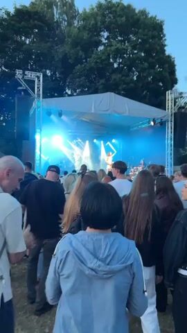 The media file shows a crowded outdoor concert in Panevėžys, Lithuania. A live band performs on a brightly lit stage, with a female vocalist center stage. The stage is covered by a white canopy and supported by metal scaffolding. Stage lights cast beams of blue and white light, illuminating the performers and creating a dynamic atmosphere. Behind the stage, tall trees are visible, suggesting the event is taking place in a park or open-air venue.

In the foreground, the backs of many concert-goers are visible, facing the stage. The crowd appears engaged and enthusiastic, with some individuals raising their hands. The time of day appears to be dusk or early evening, as the sky is dark but the stage lights are prominently featured. The overall mood is festive and energetic, typical of an outdoor music event. There is no visible text in the media file.