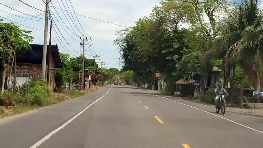 The video shows a POV shot of driving down a paved road in a rural area. The road is lined with trees, utility poles, and small buildings on either side. There are a few vehicles visible in the distance, including a yellow truck. The sky is overcast.