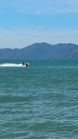 The media file captures a serene ocean scene under a clear, bright blue sky, suggesting it is daytime. In the distance, lush, green mountains form a backdrop, hinting at a coastal or island location. The sea is a vibrant turquoise, with gentle waves rippling across its surface.

The primary subject is a person riding a jet ski across the water. The jet ski is dark in color and is creating a white spray as it moves at speed. The rider is wearing dark clothing and appears to be alone on the jet ski. Their posture suggests they are actively engaged in controlling the vehicle.

The overall atmosphere is one of leisure and recreation. The bright weather and the activity of jet skiing evoke feelings of fun and enjoyment. The presence of the mountains suggests a picturesque and possibly tropical setting, consistent with the provided location context of Baan Mae Nam, Thailand. There is no visible text in the media file.