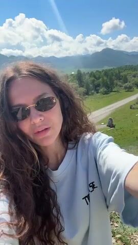 A woman with long, curly brown hair and sunglasses takes a selfie outdoors. She is wearing a white t-shirt. Behind her, a winding road runs through a grassy valley with trees and mountains in the distance. The sky is blue with scattered clouds. A white van is visible on the road.
