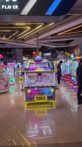 The video shows the interior of an arcade. Bright neon lights from the game machines illuminate the polished floor, creating colorful reflections. Several arcade cabinets, including claw machines and what appear to be rhythm or racing games, are visible. In the foreground, a pink and blue claw machine with a dome-shaped top and an astronaut graphic on its base is prominently featured. A few people are present in the background, some appearing to play the games, while others walk through the arcade. The ceiling has a modern design with multiple rows of yellow LED strip lights.