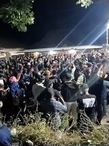 This video captures a crowded outdoor gathering at night. A large group of people, estimated to be over a hundred, are packed together, many with their arms raised. The scene is illuminated by artificial lighting, casting long shadows and creating a dynamic visual. The environment appears to be somewhat rustic, with trees and vegetation visible in the foreground. The individuals in the video are diverse in age and attire, suggesting a communal event. Some are wearing hats and casual clothing, while others are in more distinctive outfits. The overall atmosphere appears lively and energetic, with people actively engaged in the event. The camera is positioned at a high angle, looking down on the crowd.