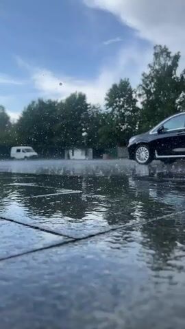 A low-angle shot shows a wet paved surface with numerous raindrops splashing. In the background, a black car is driving away from the camera on the right. Further in the background, a white van is parked on the left. The environment appears to be outdoors with trees and a cloudy sky.