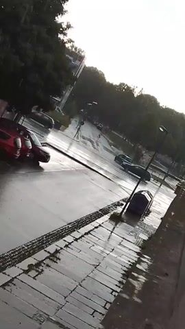 The video shows a street scene on a rainy day. The pavement and road are wet and reflective. Several cars are visible, including a red car parked on the left and other cars driving on the road. Trees line the street, and there are buildings in the background. The sky is overcast, suggesting it is daytime. The camera angle is tilted, giving a dynamic perspective of the scene.
