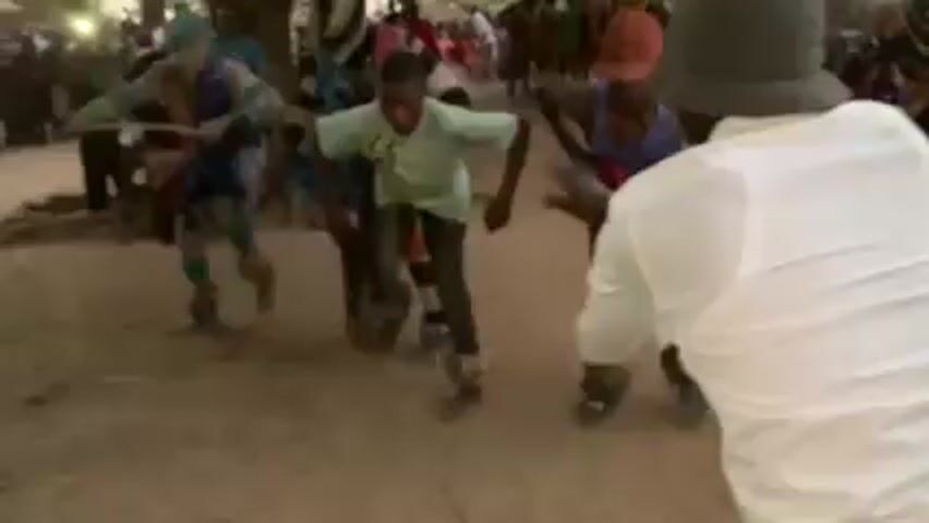 A group of dancers wearing colorful, traditional costumes perform in a dusty area. Some of the dancers have objects tied to their ankles, which create sound as they move. A person in a white shirt and a hat is positioned in the foreground, possibly filming or observing the performance. Behind the dancers, a crowd of people watches. The scene appears to be a cultural celebration taking place outdoors, likely in a village or rural setting.
