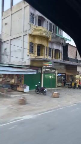 The video shows a street scene with several buildings and parked motorcycles. The buildings appear to be shops or businesses, with some displaying goods or signs. There are a few people visible, some walking or standing near the shops, and others on motorcycles. The street is paved and appears to be a busy commercial area. The video captures the general ambiance of the location as the camera pans across the scene.