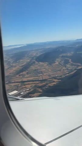 The video shows an aerial view from an airplane window. The landscape below consists of rolling hills and mountains, with patchwork fields of varying colors, suggesting agriculture. A road or river can be seen winding through the valley. The sky is a clear, bright blue. Part of the airplane's wing and window frame are visible in the foreground. The scene is illuminated by natural daylight.
