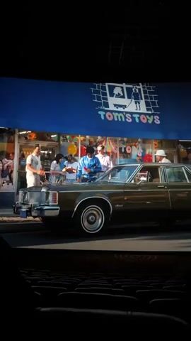 The video shows a screen in a movie theater. On screen is a clip of a movie showing a dark green car driving by. It then switches to three people standing outside a shop. A white and blue bus drives by, showing people cheering in the street. The cheering people are blurred.