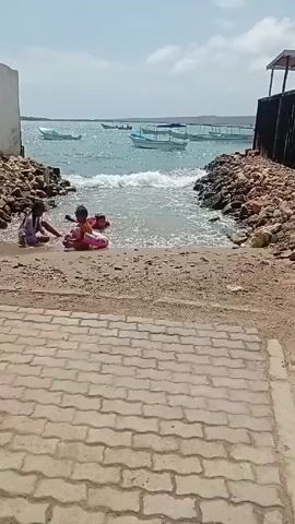 The scene is a beach inlet on a sunny day, with a blue and partly cloudy sky. Two children are playing in the shallow water where the inlet meets the ocean. One child is wearing a ring-shaped floatation device. Boats are visible in the background on the water, a pier is visible on the right. The beach is a mix of sand and rocks, with brick-paved steps leading down to the water.  The inlet is bordered by rocks on both sides, and a small building structure is on the far right. The ocean has small waves, and the water near the shore is murky.