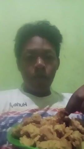 A person is holding a green plate full of fried chicken pieces. The person picks up a piece of chicken with their right hand and brings it to their mouth, then chews and swallows. The person is wearing a white t-shirt with a colorful pattern. The background is a plain green wall.