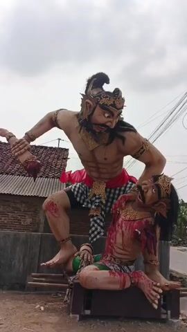 The video shows two large, elaborate statues, likely figures from a myth or folktale, depicted in a violent scene. One statue, standing over the other, is much larger and appears to be male, with a muscular physique, dark hair, and ornate gold headwear. This figure is raising a weapon, possibly a club or mace, in its right hand, and its left hand is gripping the head of the smaller statue. The smaller statue is kneeling and appears to be in distress or defeat. It also has a dark mustache and ornate gold headwear. Both statues are covered in what appears to be fake blood, suggesting a violent encounter. The standing figure has red markings on its legs, and the kneeling figure has red markings on its torso and arms, indicating wounds. The statues are positioned outdoors, with a dusty ground, a simple building with a tiled roof in the background, and utility poles with wires to the right. The sky is overcast. The scene is staged and appears to be part of a cultural display or performance.