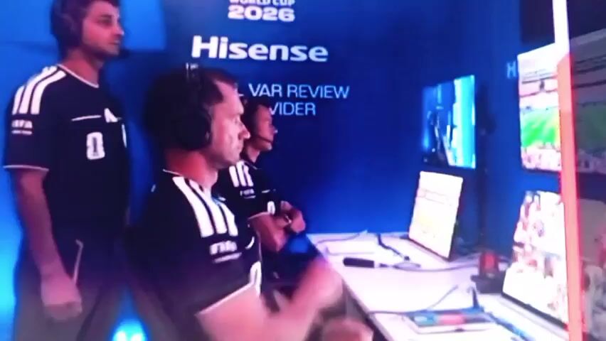 The video shows three individuals, two seated and one standing, in what appears to be a VAR (Video Assistant Referee) room. The individuals are wearing black shirts with white stripes on the sleeves and FIFA logos. The standing individual on the left is wearing headphones and looking at a screen. The two seated individuals are also wearing headphones. The individual in the foreground on the left is seated and has their arms crossed, looking at a screen in front of them. Behind them, another individual is seated and also looking at a screen. The room is well-lit with blue lighting. The background displays "WORLD CUP 2026" and "Hisense OFFICIAL VAR REVIEW PROVIDER". Multiple screens are visible, showing various game footage and data. One screen shows a close-up of a soccer game. Another screen appears to be displaying tactical information or statistics. The overall impression is one of focused attention and technical operation within a professional sports context.