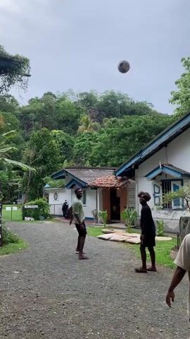 The video shows three men playing keep-ups with a brown coconut outside a white building with a blue-trimmed roof. The men are dressed casually. One wears a black top and shorts, another a green shirt and black shorts, and the third a beige shirt. The building has blue shutters on the windows and is surrounded by trees and greenery. The ground is covered in gravel. A sign with foreign characters can be seen in the background. The coconut is thrown and kicked between the men as they try to keep it in the air.