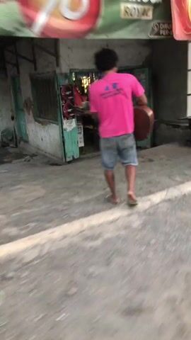 The media file shows a person carrying a guitar while walking on a street. The person is wearing a bright pink t-shirt and denim shorts, with flip-flops on their feet. The guitar is slung over their shoulder, held by a strap.

The scene appears to be outdoors, on a paved or dirt road, with buildings visible in the background. The lighting suggests it is daytime, possibly late afternoon or early evening, given the softer light and shadows. The general atmosphere seems casual, with the person likely engaged in a personal activity, such as going to or from a musical performance or practice.

The location context provided is Kebakalan, Indonesia. This suggests a potentially urban or semi-urban setting in Indonesia.

On the back of the person's pink t-shirt, there is visible text. Although blurred, it appears to be a logo or name with several lines of text. The first line seems to be "AR" followed by more text. The second line also contains text, and a third line is present below that. The text is in a sans-serif font and appears to be in white or a light color against the pink fabric.

The ground is uneven and dusty, typical of a street in a less-developed area. The background shows a building with some shelves or displays, and other structures that are out of focus. The overall image quality is low, with a significant amount of motion blur, making it difficult to discern finer details of the surroundings or the person's facial features.