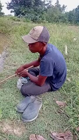 A person wearing a blue t-shirt and a cap sits on the grass by the edge of a muddy river, holding a fishing rod. They appear to be actively fishing, with the rod extended over the water. A plastic bag and a pair of rubber shoes are on the ground near them. The environment is rural, with lush green vegetation and trees lining the riverbanks. The camera pans slightly to show more of the river and its surroundings, with a focus on the fishing activity.