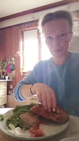 A woman with curly hair and a light blue sweatshirt is sitting at a table with a plate of food. The food on the plate includes a piece of grilled fish, some greens, and a pastry. The woman is using a fork to cut into the pastry. She is looking at the camera and smiling. The background of the scene is a kitchen with wooden cabinets and a window with bright sunlight coming through.