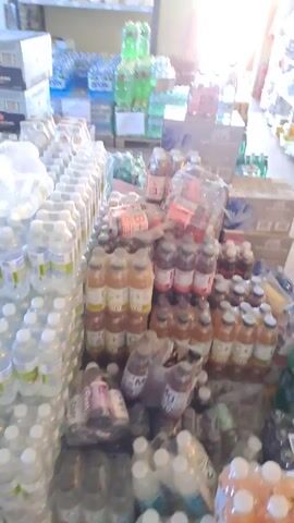 The video pans across a room filled with shelves and stacked cases of drinks. The shelves on the left are stocked with bottles of water and other beverages, while the shelves on the right hold various packaged goods. In the foreground and middle of the room, there are numerous cases of bottled drinks, including soda and juice. The lighting appears to be bright, suggesting the presence of natural light from a doorway or window at the end of the room. The room appears to be a storage area or a retail space.