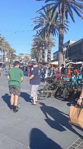 The video shows a pedestrian area lined with palm trees and buildings on a sunny day. People are walking around and sitting on benches. There are several bicycles parked along the street, and an outdoor dining area is visible at one of the restaurants. Two men are standing and talking in the foreground, and one of them is holding a bottle of water.