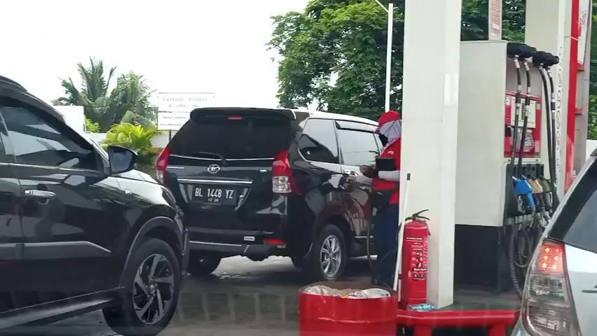 The video shows a gas station scene with several vehicles lined up. In the foreground, a black SUV is partially visible on the left. Behind it, a black minivan with license plate DL 1448 YZ is parked at a gas pump. A person wearing a red and white uniform is attending to the black minivan, appearing to be refueling it. A red fire extinguisher is mounted on the gas pump island. In the background, there are trees and a sign with text. The weather appears overcast. The video captures a typical moment of vehicles being serviced at a gas station.