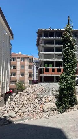 The video captures an outdoor scene on a clear, sunny day in Bahçelievler, Turkey. The primary subjects are a multi-story building under construction and a pile of demolition debris in the foreground. The building is a concrete structure with scaffolding and safety netting visible. Several figures, presumably construction workers, are present on the top level of the unfinished building.

Adjacent to the construction site, there are existing apartment buildings, suggesting an urban or semi-urban setting. A tall, overgrown vine-covered pole stands prominently to the right of the construction site, contrasting with the man-made structures. The debris pile consists of concrete, bricks, and other construction remnants. A white car is parked behind the debris pile, and other vehicles are visible in the background.

The time of day appears to be daytime, given the bright sunlight and clear blue sky. The weather seems fair. The overall impression is one of ongoing construction and development, juxtaposed with remnants of past structures. No visible text is present in the video.