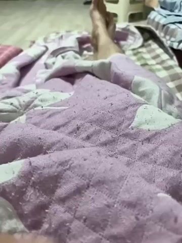 The video shows a close-up view of a person's legs under a lavender and white patterned blanket. The blanket appears to be made of a soft, textured fabric, possibly cotton or a blend, with a quilted diamond pattern. There are small pills or fuzz balls visible on the surface of the blanket, suggesting it might be well-used.

In the background, slightly out of focus, a person's feet are visible. They are partially covered by another blanket or sheet with a checkered pattern of brown and white. The overall setting appears to be indoors, possibly a bedroom, given the presence of blankets and what looks like a bed. The lighting suggests it might be daytime or a well-lit indoor space, with no clear indication of natural light sources.

The activity taking place is subtle. A hand, adorned with a ring on the index finger, enters the frame from the lower left and gently touches and pulls at the lavender blanket. The fingers are pale and manicured. The interaction with the blanket is gentle, perhaps indicating a moment of relaxation, comfort, or simply an exploration of the texture.

There is no visible text in the video. The mood is calm and intimate, focusing on the tactile experience of the blanket and the quiet presence of the person. The location context of Sejong, South Korea, doesn't offer specific visual cues within this close-up shot, but it places the scene within a particular geographical and cultural setting. The time of day is indeterminate.