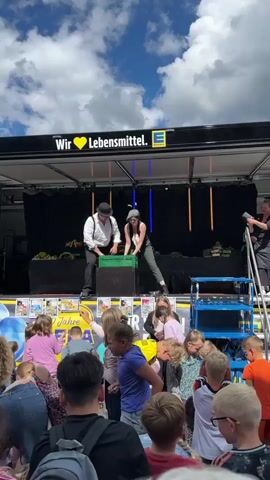 A performance is taking place on a raised stage in front of a crowd of people, mostly children. The stage is set up on the back of a truck or trailer with a black backdrop and a banner that reads "Wir ♥ Lebensmittel." and the Edeka logo. Two performers are on the stage, a man and a woman. The man is wearing a white shirt, suspenders, black pants, and a hat. The woman is wearing a dark vest, a light-colored shirt, and patterned pants, with a hat. They are interacting with a green crate. The man lifts the crate, and the woman appears to be holding it. In the foreground, a crowd of children and adults are watching the performance. Some children are closer to the stage, reaching out. The environment appears to be outdoors, with a cloudy blue sky overhead. Balloons, some yellow and gold, are visible on the left side of the stage. There are also posters and advertisements visible on the front of the stage.