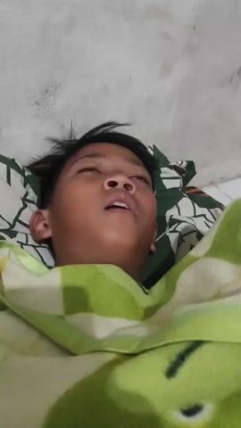 The video shows a young person lying in bed, covered by a blanket. The person's eyes are closed, and their mouth is slightly open, suggesting they are asleep or resting. The person is lying on a pillow with a green and white pattern. The background is a plain, light-colored wall.