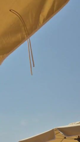 The video shows a low-angle shot of a clear blue sky. Two beach umbrellas are partially visible at the bottom of the frame. The umbrella in the foreground is a light yellow fabric with a white edge. A string hangs from it. Another umbrella, also light yellow with a white edge, is visible behind and to the right of the first one. The top of the frame shows a close-up of a yellow beach umbrella. The video is static.