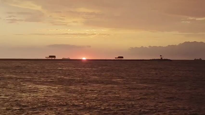 A drone shot captures a serene ocean at sunset. The sun, a vibrant orange orb, is low on the horizon, casting a warm glow across the sky and water. Two small, stilted structures stand on a distant pier, silhouetted against the twilight. The water is a deep, muted brown, with gentle ripples reflecting the sky's colors. A distant ship is visible on the left, and a lighthouse on the right. The clouds, streaked with orange and yellow, add to the tranquil atmosphere.