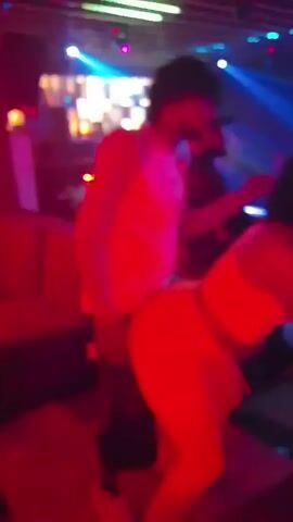The video shows two people dancing in a club with colorful lighting. A person with curly hair wearing a white tank top and dark pants dances behind a person wearing a white tank top and pants. A DJ wearing a baseball cap stands in the background.