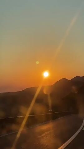 This video captures a scenic drive along a winding road in Markovići, Montenegro, during a breathtaking sunset. The primary subject is the setting sun, a brilliant orb of light casting a warm, golden glow across the sky and the surrounding landscape. Rays of sunlight streak across the frame, creating a dreamy and ethereal atmosphere.

The scene is dominated by rolling hills and mountains, silhouetted against the vibrant hues of the sunset. A road, marked with white lines, curves through the foreground, suggesting movement and a journey. A red car is visible on the road, adding a sense of human presence and activity to the otherwise natural panorama. The road appears to be a mountain pass, with a guardrail indicating a drop-off, likely a scenic overlook or a route with stunning vistas.

The time of day is clearly sunset, evident from the low angle of the sun and the warm, orange and yellow tones in the sky. The weather appears clear, allowing for an unobstructed view of the sunset. The location cues, specifically "Markovići, Montenegro," place the scene in a mountainous coastal region known for its natural beauty.

The overall emotion evoked by the video is one of peace, beauty, and perhaps a sense of wanderlust. The natural spectacle of the sunset, combined with the imagery of a road trip, creates a feeling of serenity and awe. There is no visible text in the video.